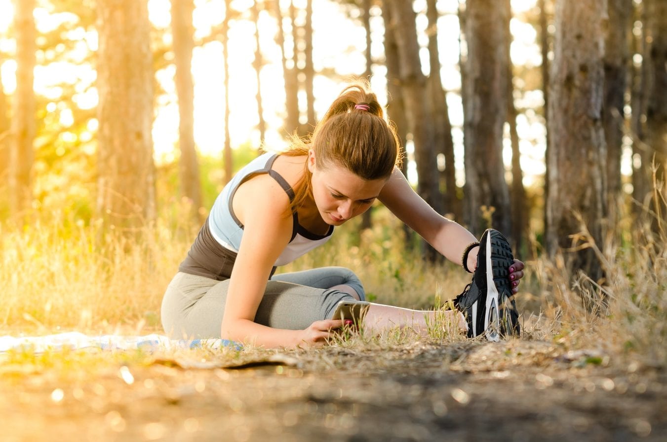 a woman is sitting on a yoga mat in the woods stretching her legs .