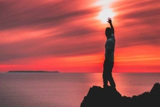 a man is standing on top of a rock overlooking the ocean at sunset .