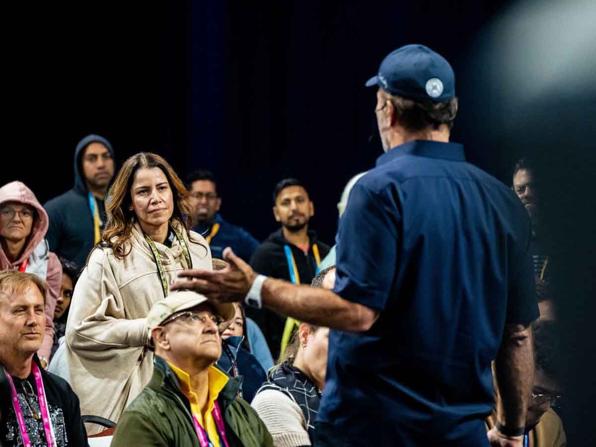 A man in a blue shirt and cap speaks to a diverse audience, with a woman in a beige coat listening intently.