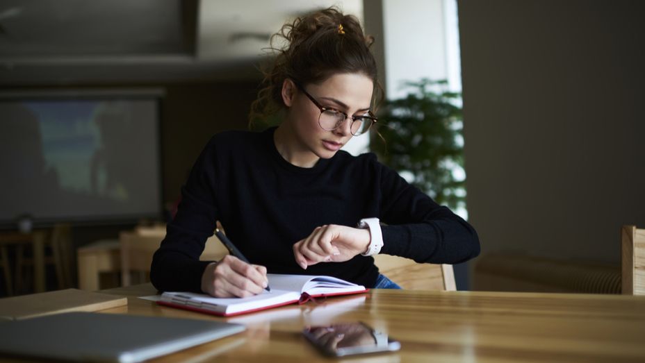 a woman is sitting at a table writing in a notebook and looking at her watch .