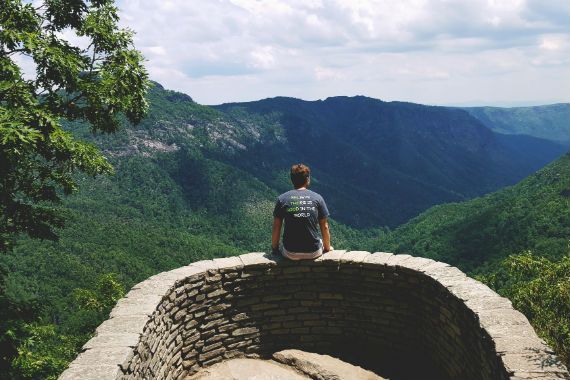 a man is sitting on a stone wall overlooking a mountain range .
