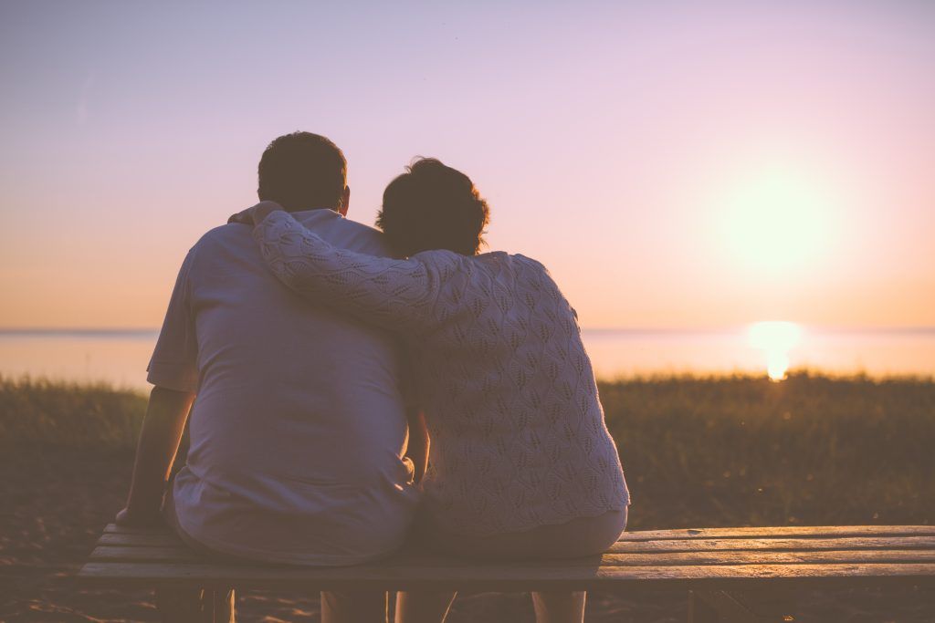 a man and a woman are sitting on a bench watching the sunset .