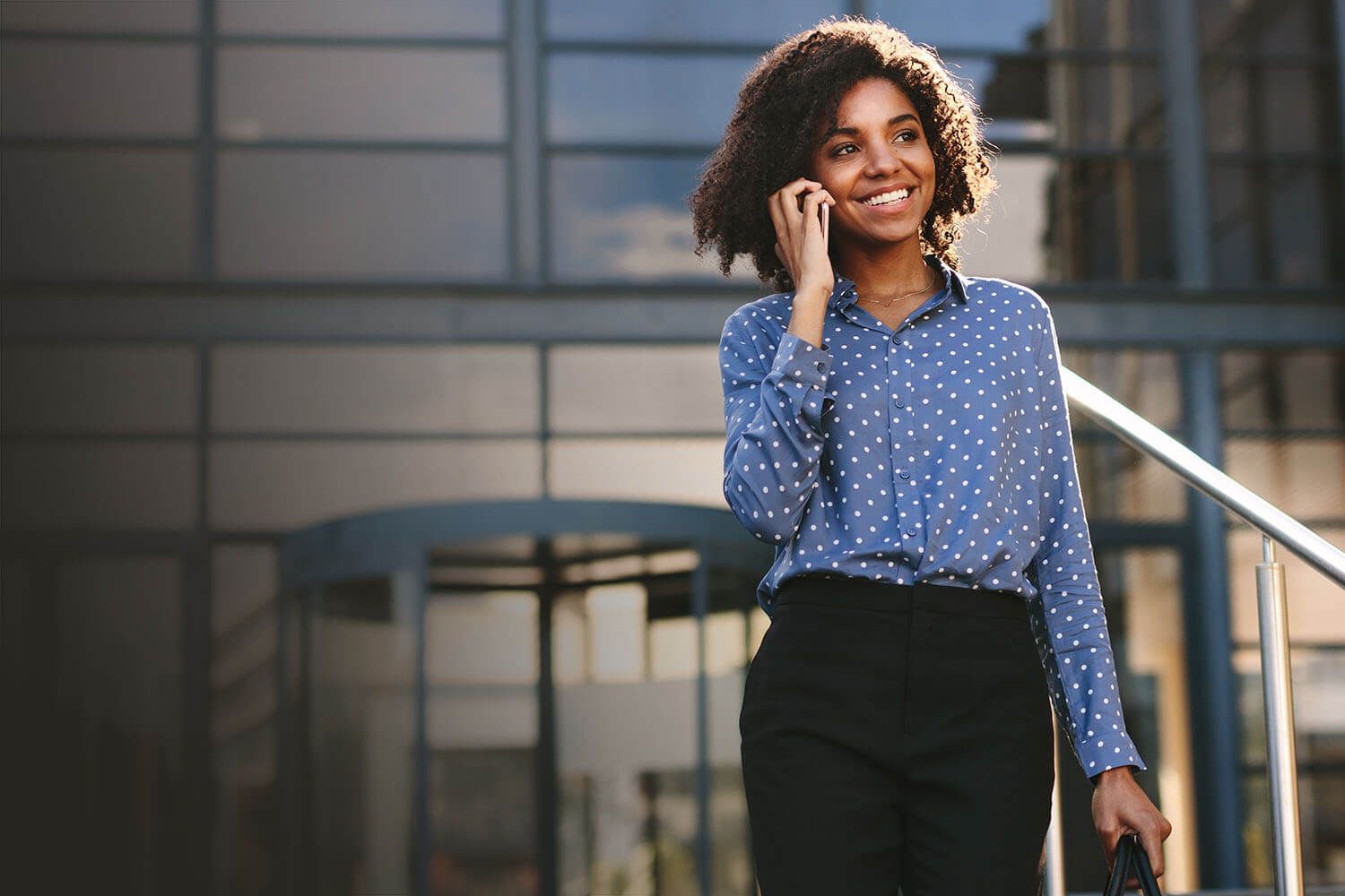 A smiling Black woman with curly hair talks on a phone while walking outside a modern building.