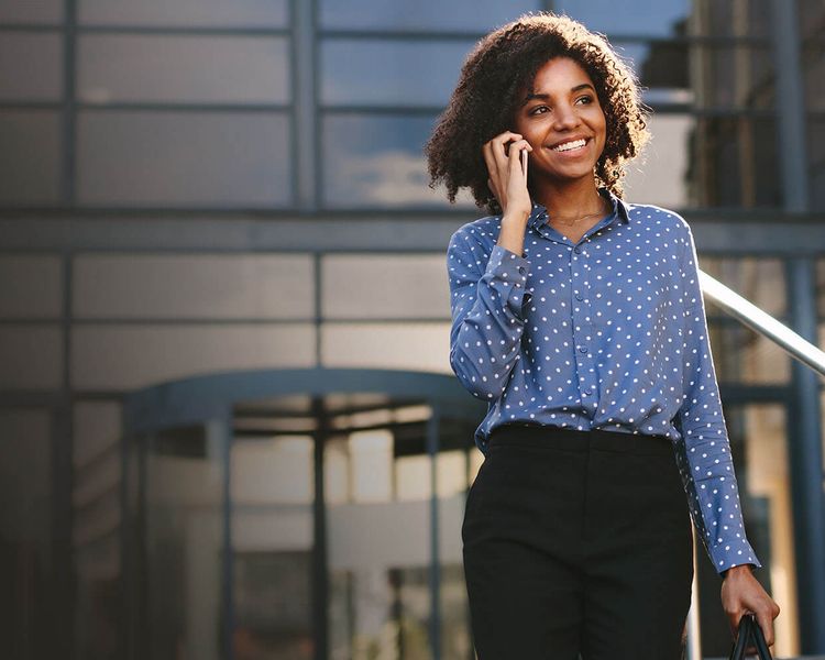 A smiling Black woman with curly hair talks on a phone while walking outside a modern building.