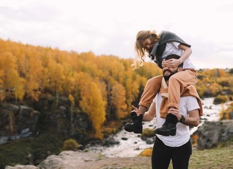 A woman riding on a man's shoulders playfully covers his eyes, with yellow autumn trees and a river in the background.