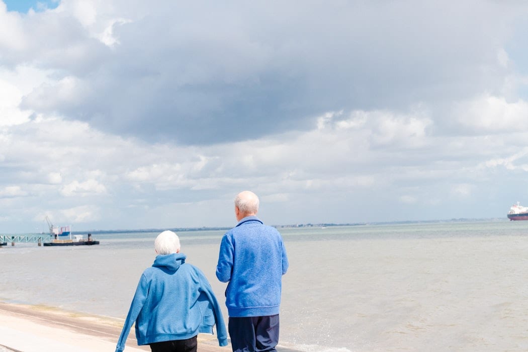 an elderly couple is walking along the beach holding hands .
