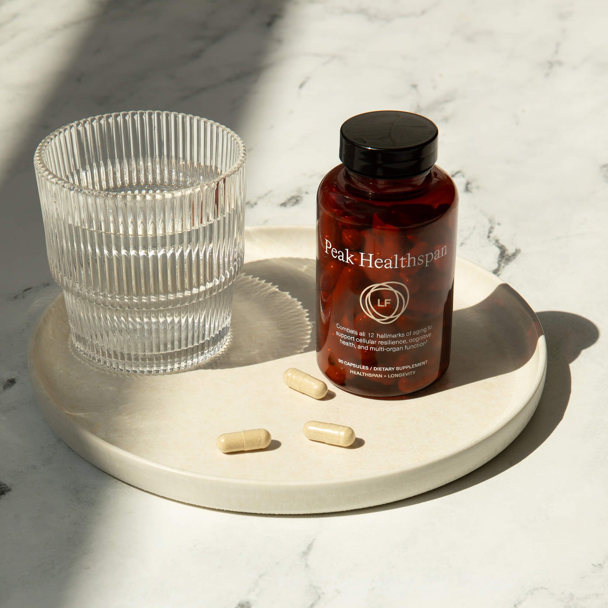 An amber bottle of Peak Healthspan supplement, a glass of water, and three beige capsules on a light tray.
