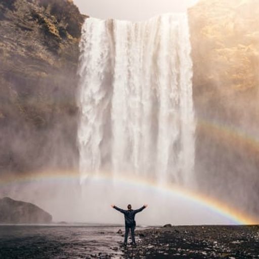 a man is standing in front of a waterfall with a rainbow in the background .