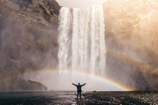 a man is standing in front of a waterfall with a rainbow in the background .