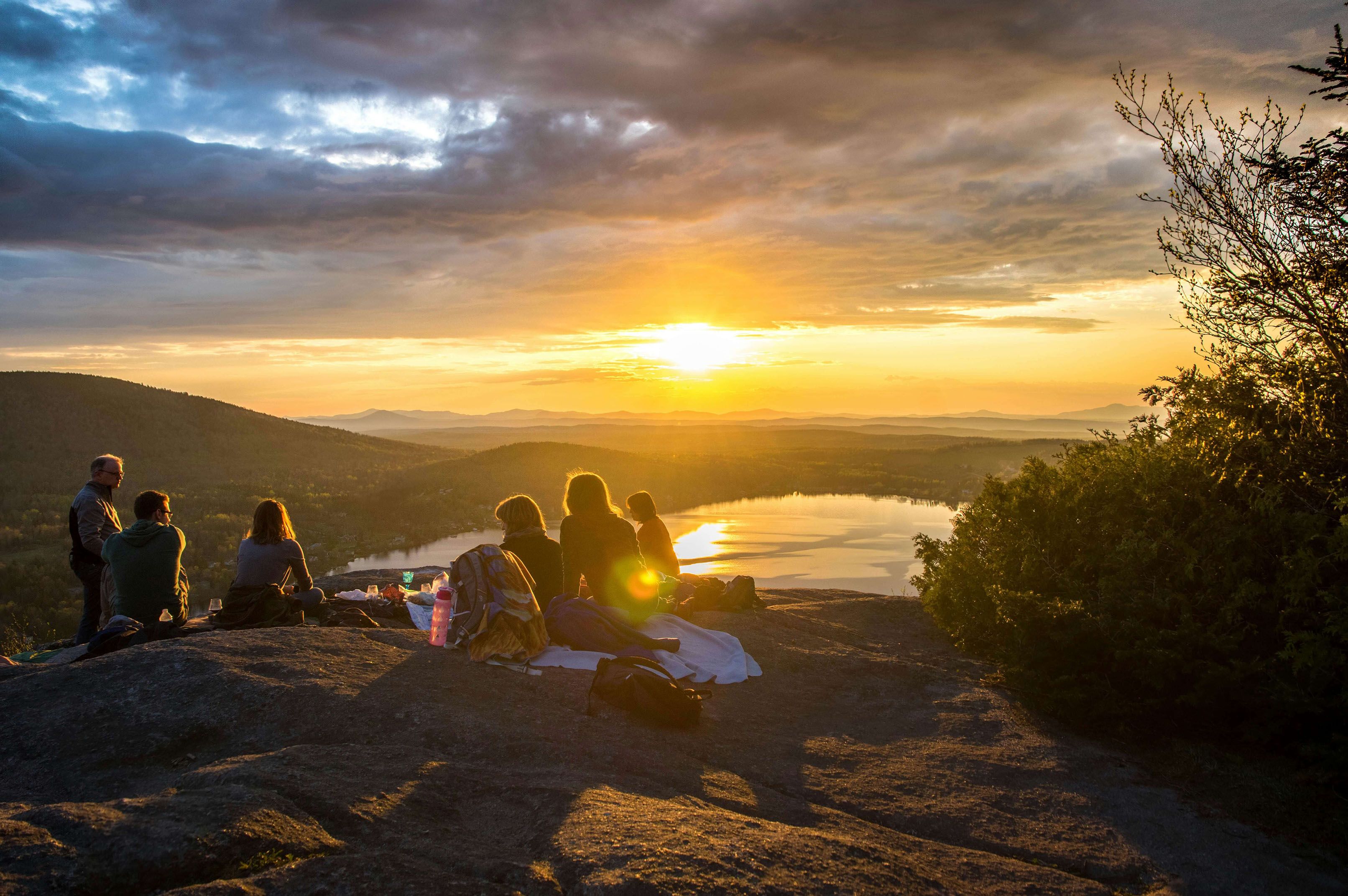a group of people are sitting on top of a rock watching the sunset over a lake .