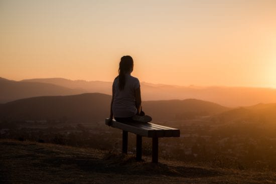 a woman is sitting on a bench looking at the sunset over the mountains .