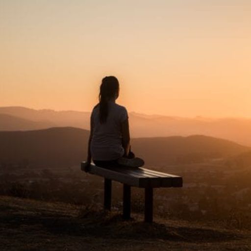 a woman is sitting on a bench looking at the sunset over the mountains .
