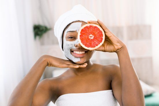 a woman with a clay mask on her face is holding a grapefruit in front of her eye .