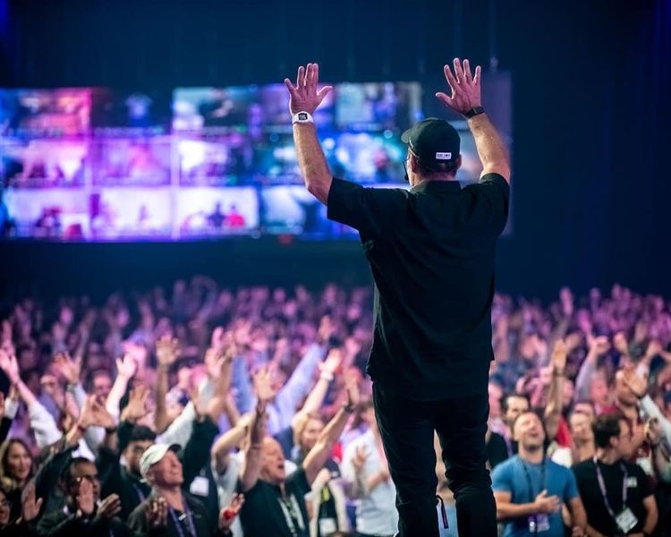 A speaker on stage with arms raised, facing a large audience with raised hands, under screens showing multiple video feeds.