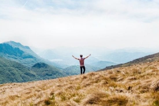 a man is standing on top of a grassy hill with his arms outstretched .