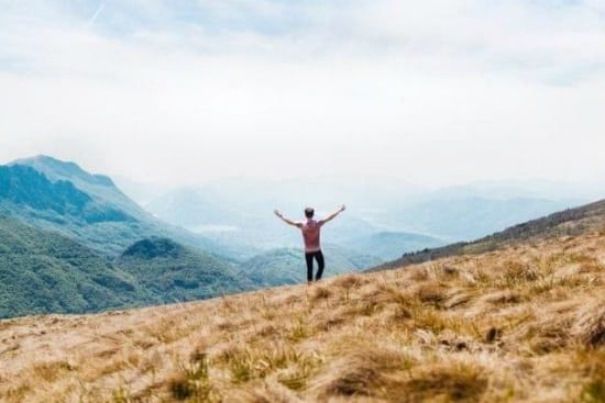 a man is standing on top of a grassy hill with his arms outstretched .