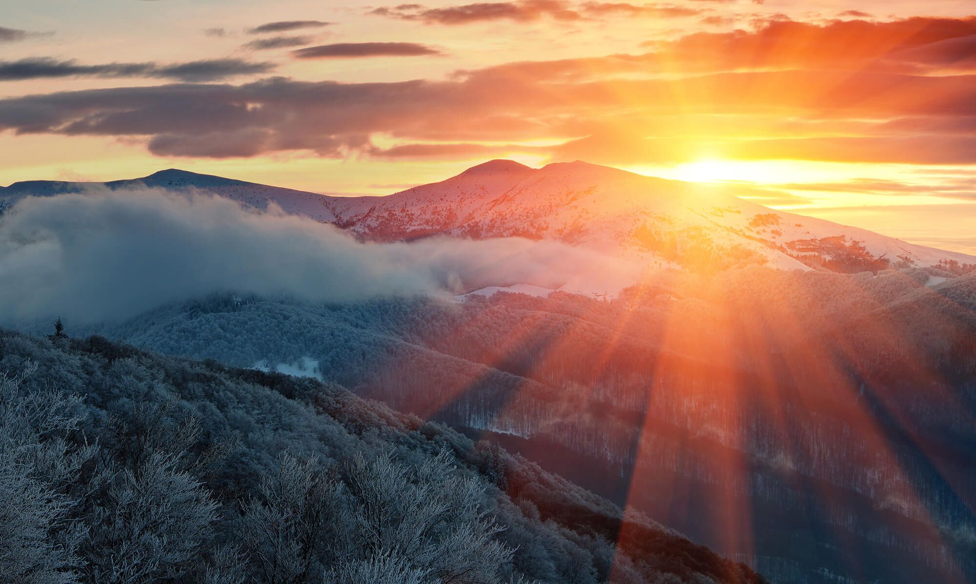 Vibrant sunset with sun rays over snow-covered mountains and frosted trees.