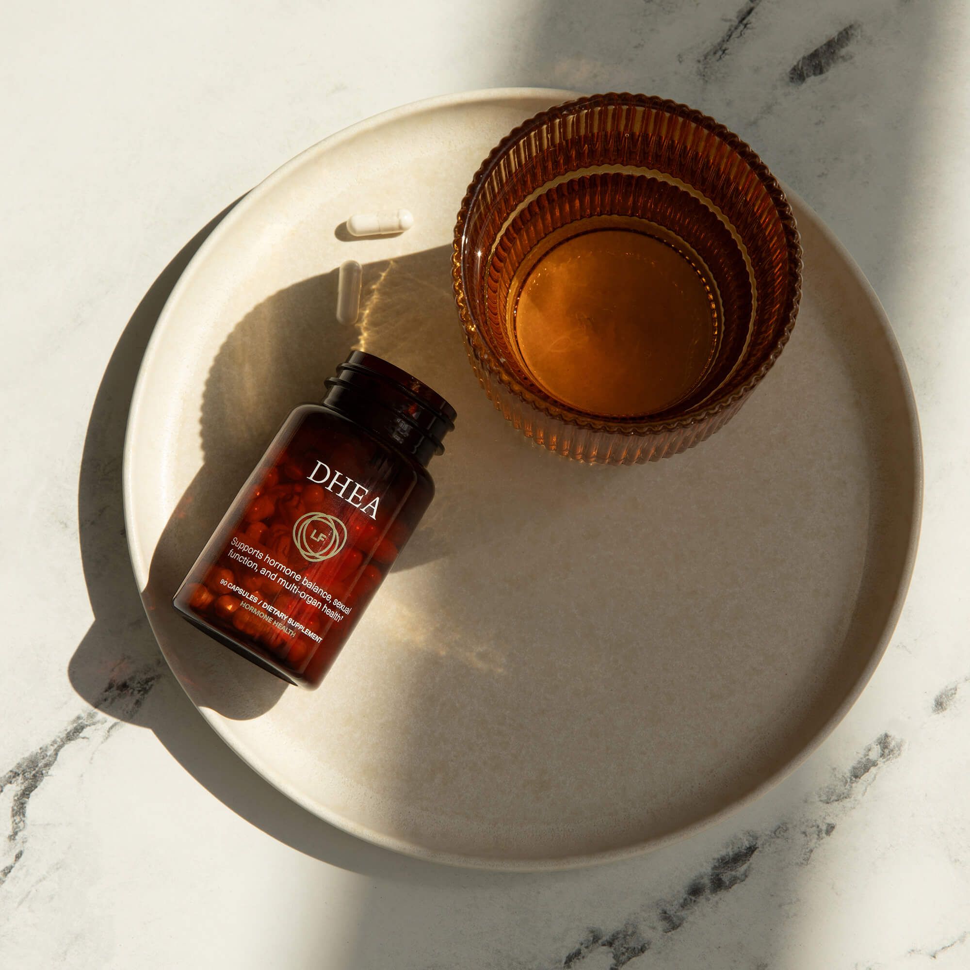 A bottle of DHEA capsules, a single capsule, and an amber glass on a round plate, against a marble background.