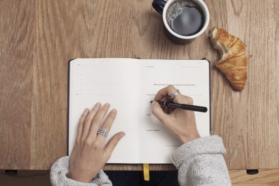 a person is writing in a notebook next to a cup of coffee and a croissant .