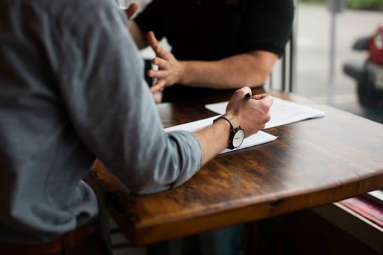 two men are sitting at a wooden table having a conversation .