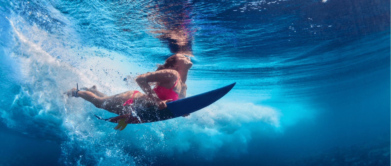 Underwater, a woman in a pink bikini holds her surfboard, looking up at a breaking wave.