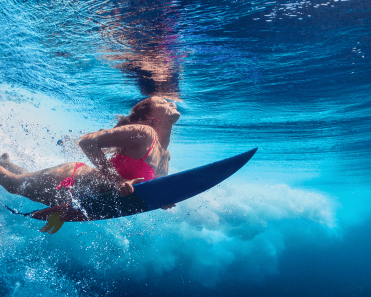 Underwater, a woman in a pink bikini holds her surfboard, looking up at a breaking wave.