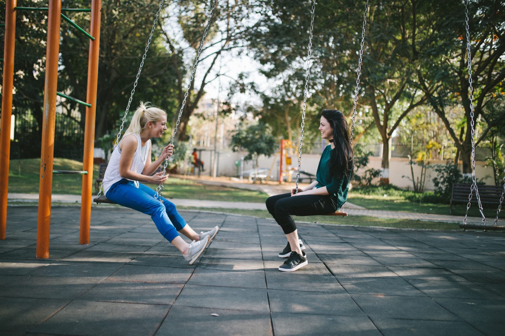 two women are sitting on swings in a park talking to each other .