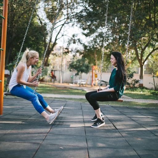 two women are sitting on swings in a park talking to each other .