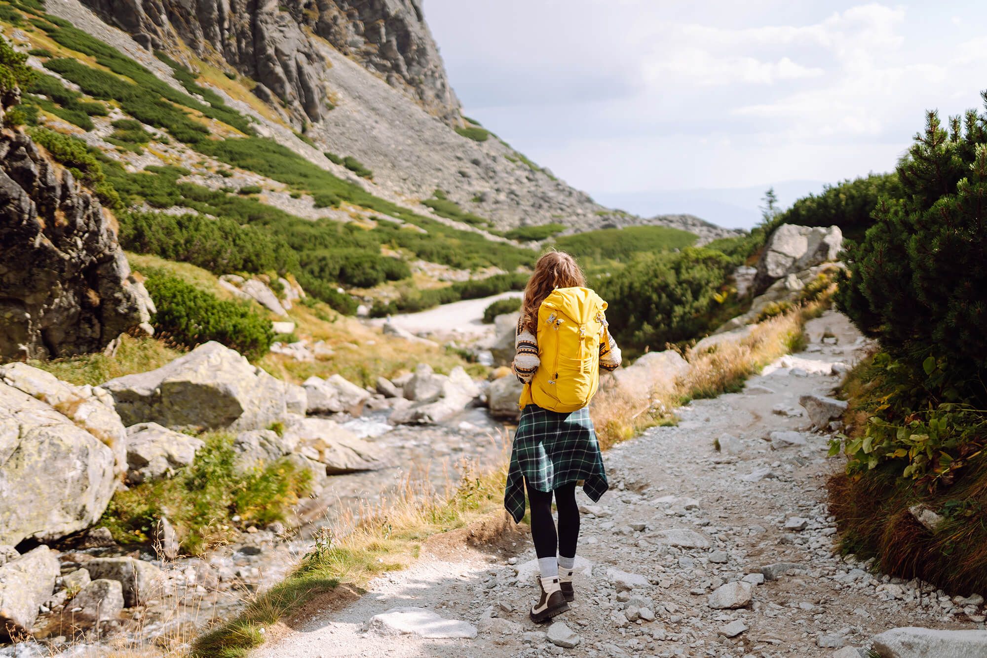A person with a yellow backpack hikes on a rocky mountain path beside a stream.
