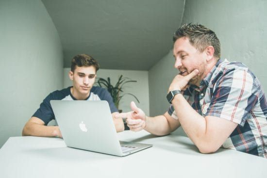 two men are sitting at a table looking at a laptop computer .