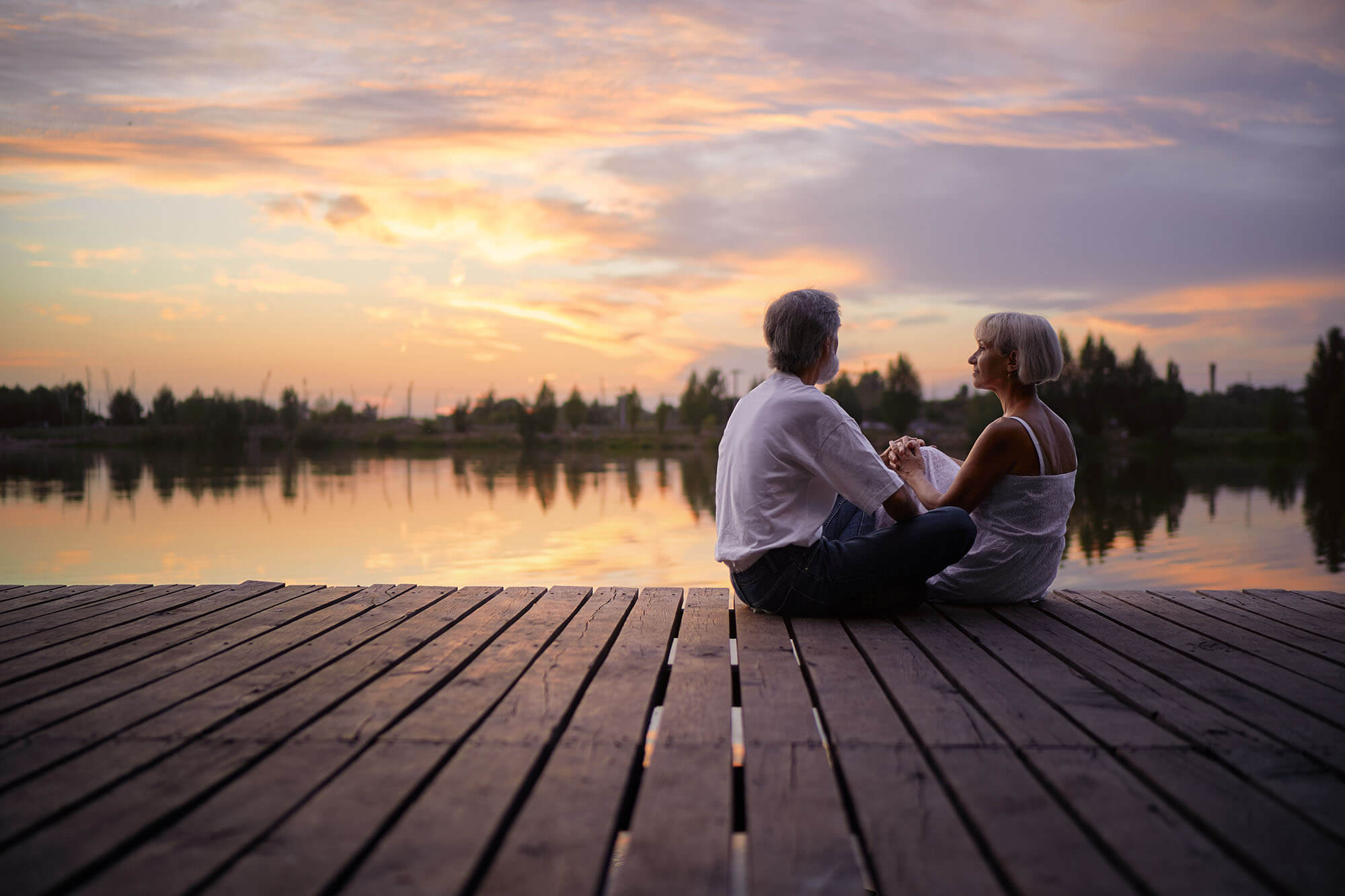 Senior couple holding hands on a dock by a lake at sunset.