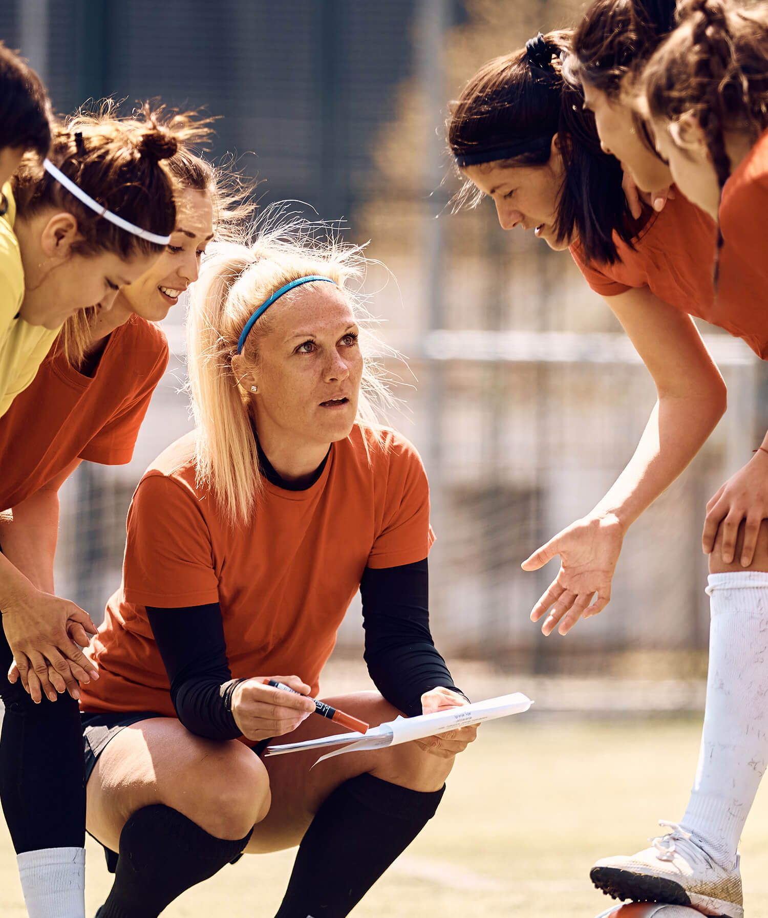 A female soccer coach crouches on the field, holding a clipboard and pen, while huddling with her team of female players.
