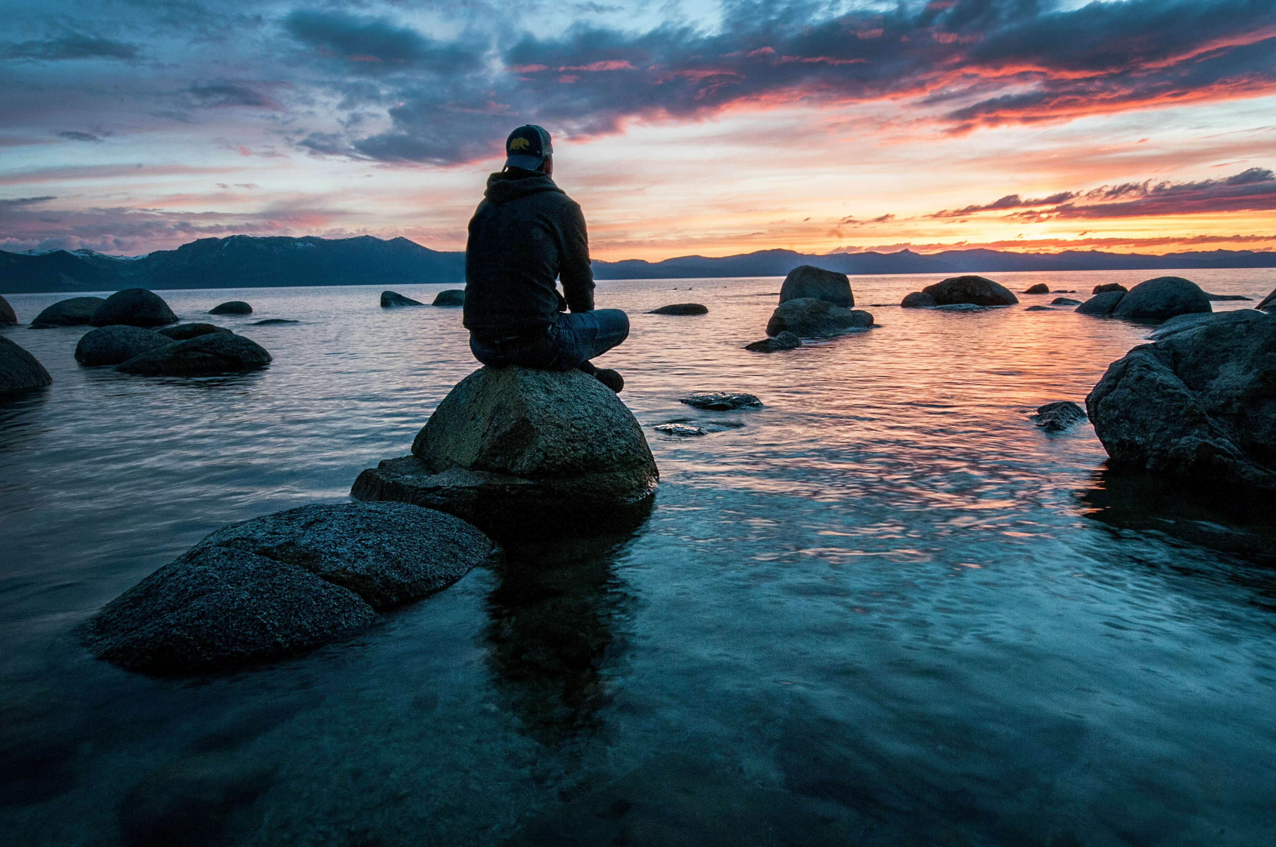 a man is sitting on a rock overlooking a lake at sunset .