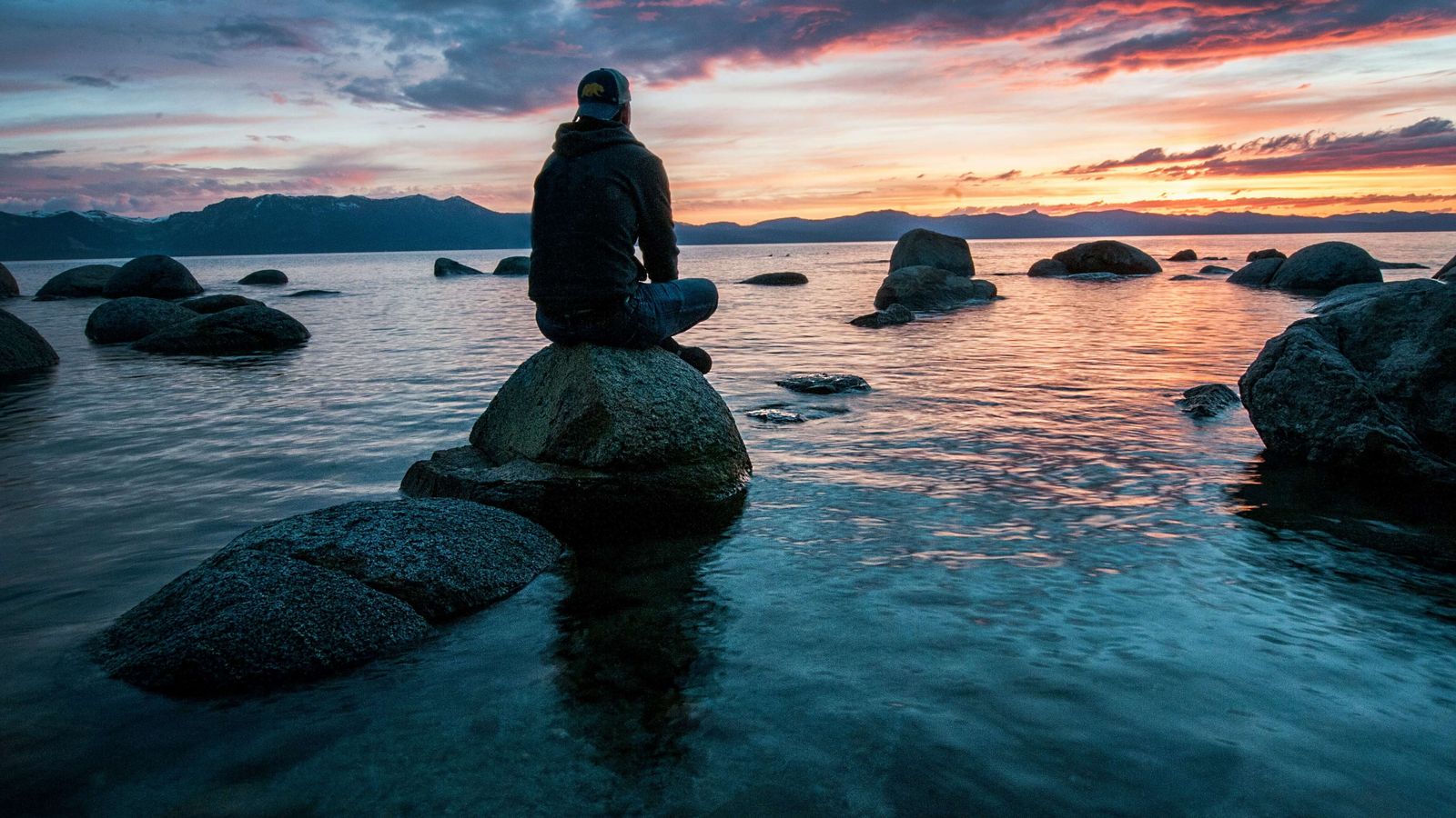 a man is sitting on a rock overlooking a lake at sunset .