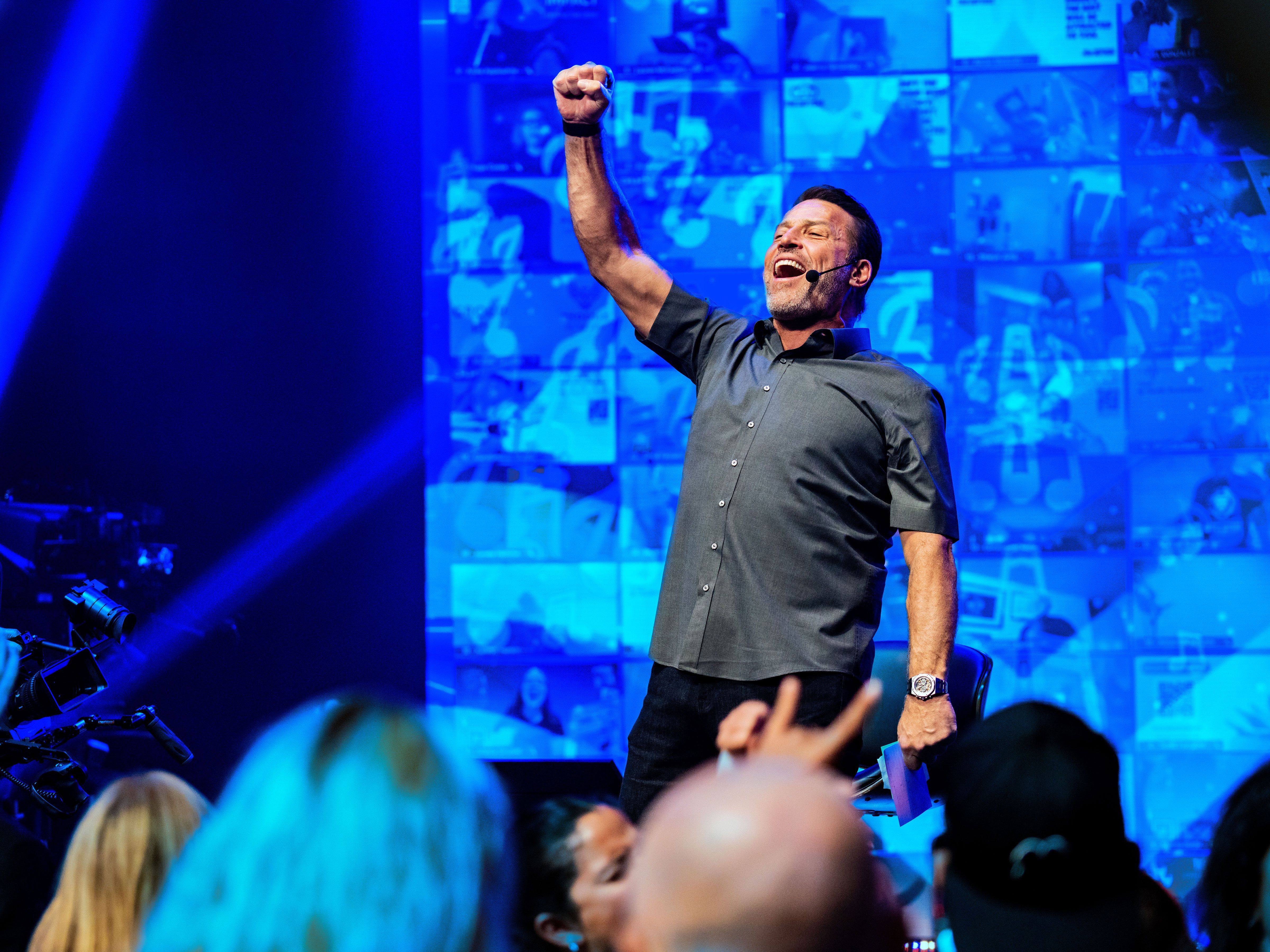 Tony Robbins triumphantly raising his arm on a blue-lit stage with screens and an audience.