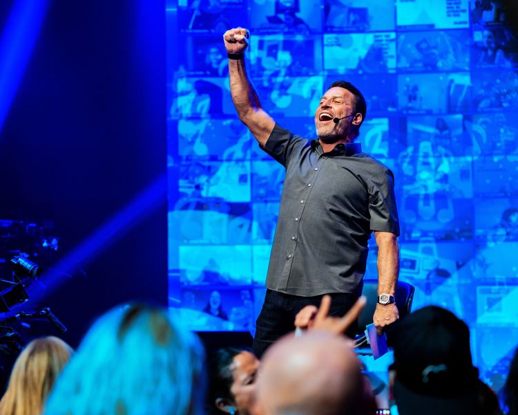 Tony Robbins triumphantly raising his arm on a blue-lit stage with screens and an audience.
