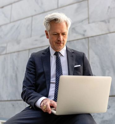 a man in a suit and tie is sitting on a bench using a laptop computer .
