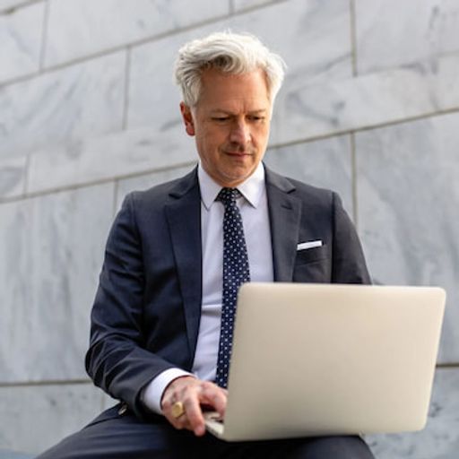a man in a suit and tie is sitting on a bench using a laptop computer .