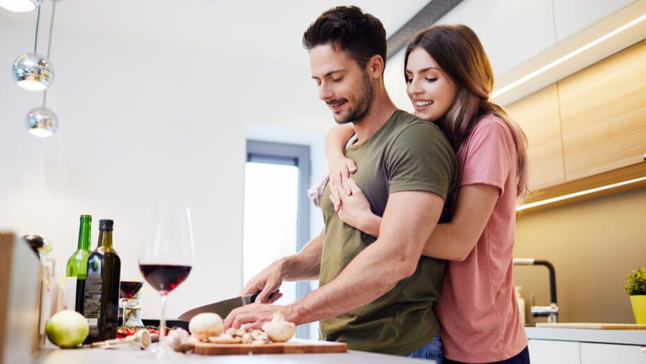 a woman is hugging a man while he cuts vegetables in the kitchen .