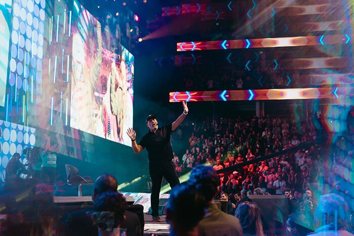A man on a stage with raised arms, facing a large audience under bright lights and colorful digital displays.