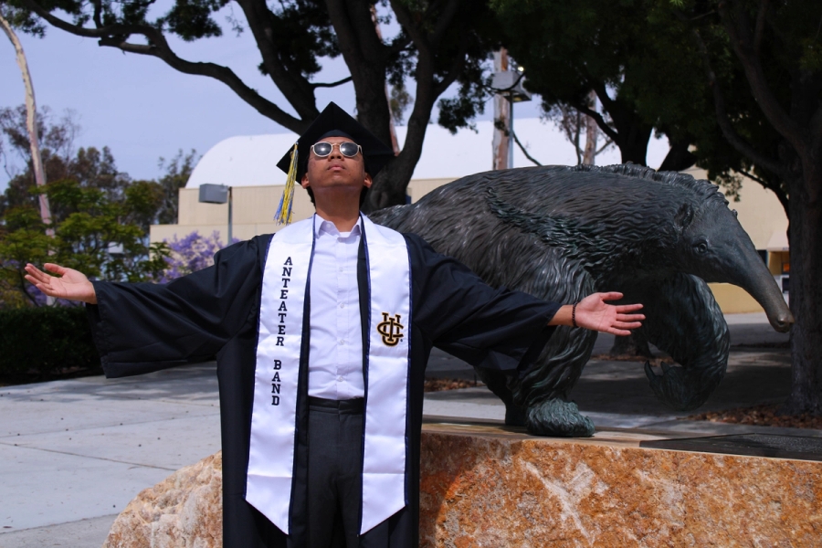 Boy in grad fit in front of anteater statue with arms up.