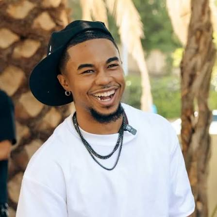 DeenTheGreat smiling in a white T-shirt, black backwards cap and layered chain necklaces outdoors