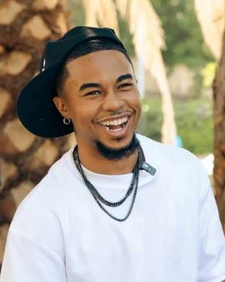 DeenTheGreat smiling in a white T-shirt, black backwards cap and layered chain necklaces outdoors
