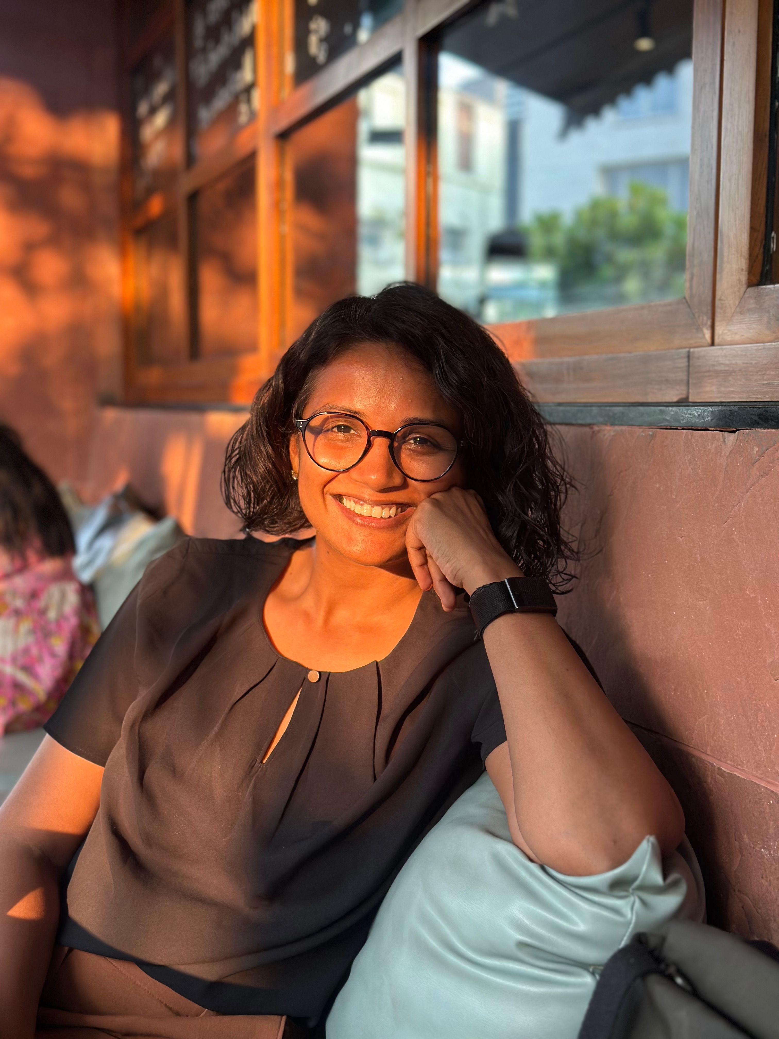 A smiling woman with glasses and dark curly hair, bathed in warm orange light, leaning on a pillow.