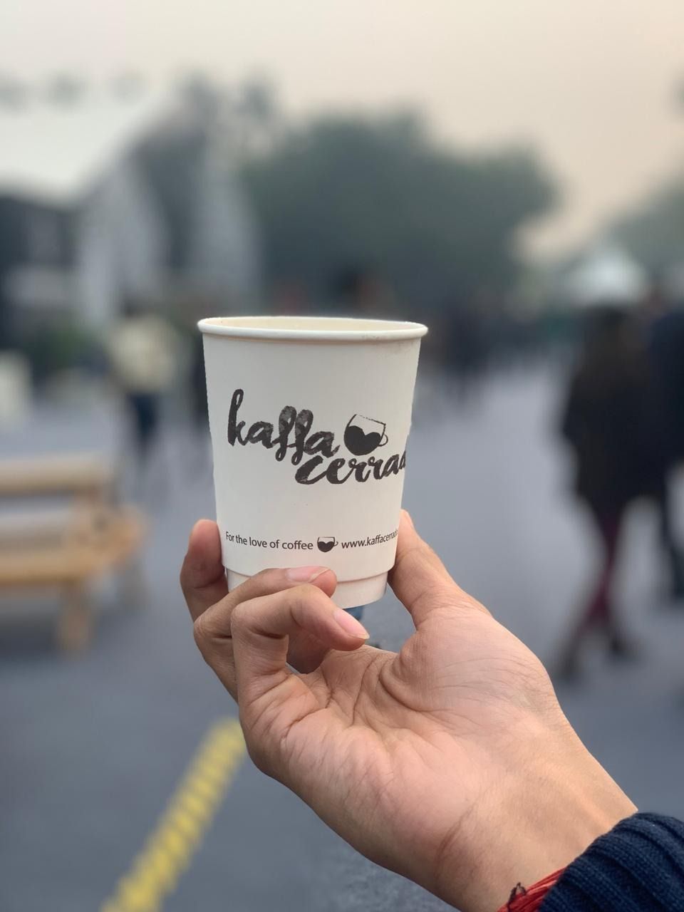 Hand holding a white "Kaffa Cerrada" coffee cup in a blurred outdoor setting.