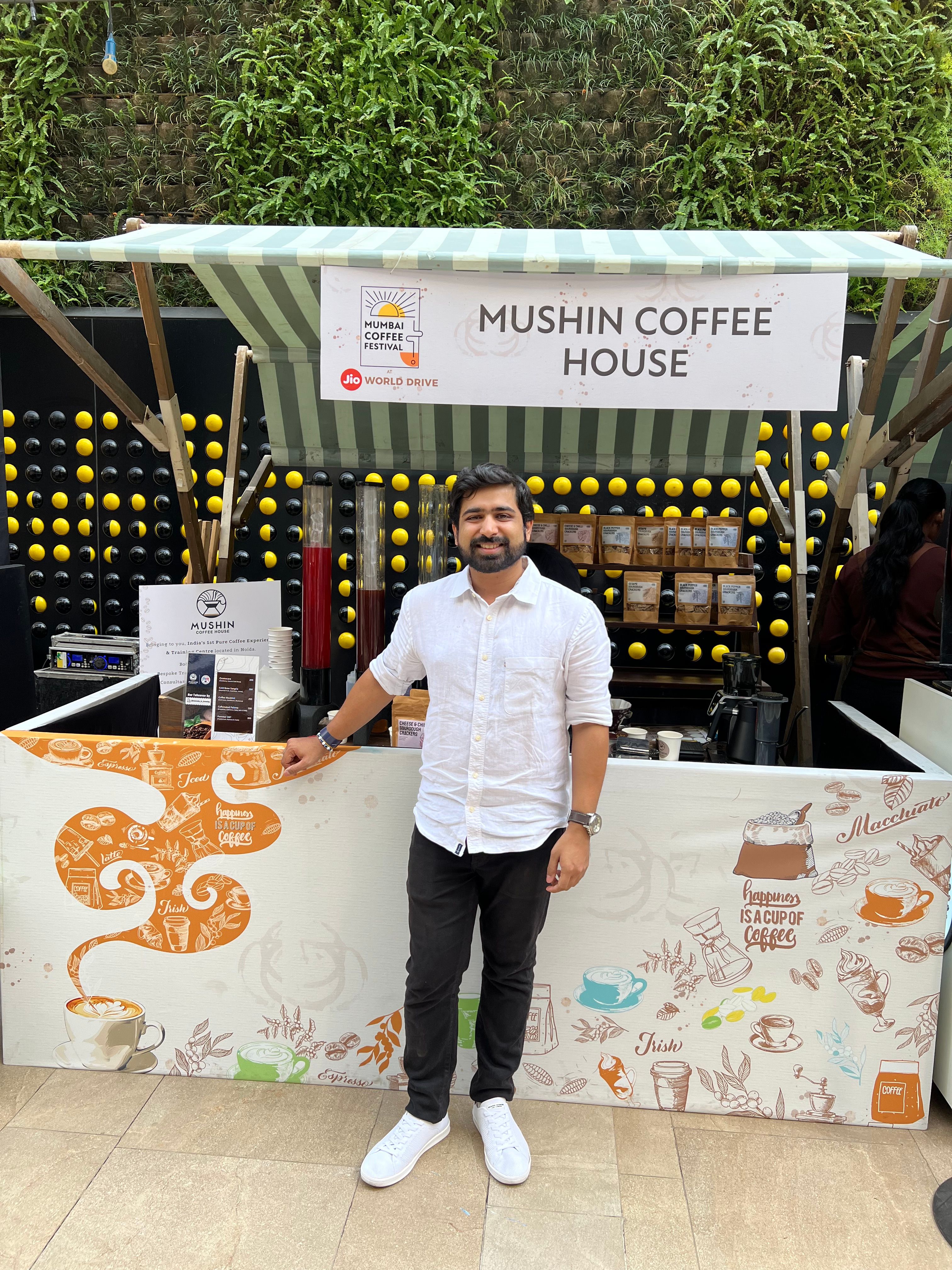 A smiling man stands in front of a Mushin Coffee House stall at the Mumbai Coffee Festival, with coffee products displayed and a green plant wall behind him.