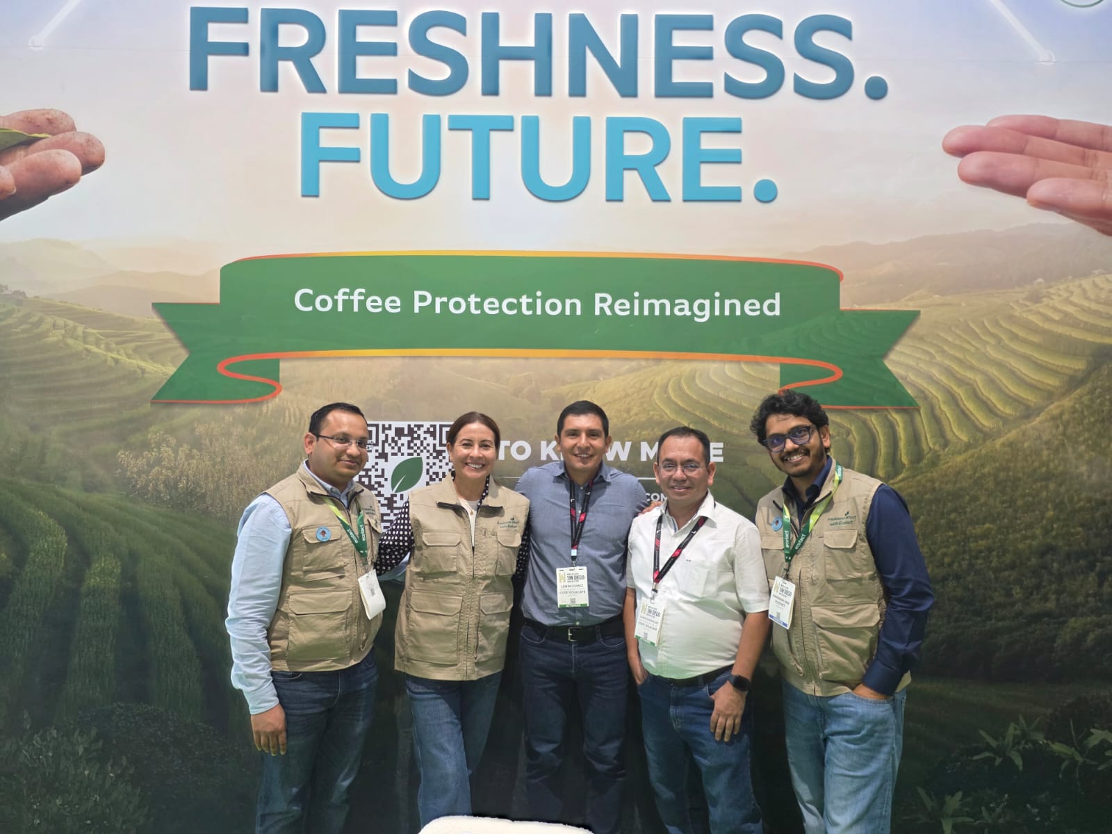 Five people, including one woman, stand in front of a banner featuring terraced coffee fields and text reading "FRESHNESS. FUTURE." and "Coffee Protection Reimagined".