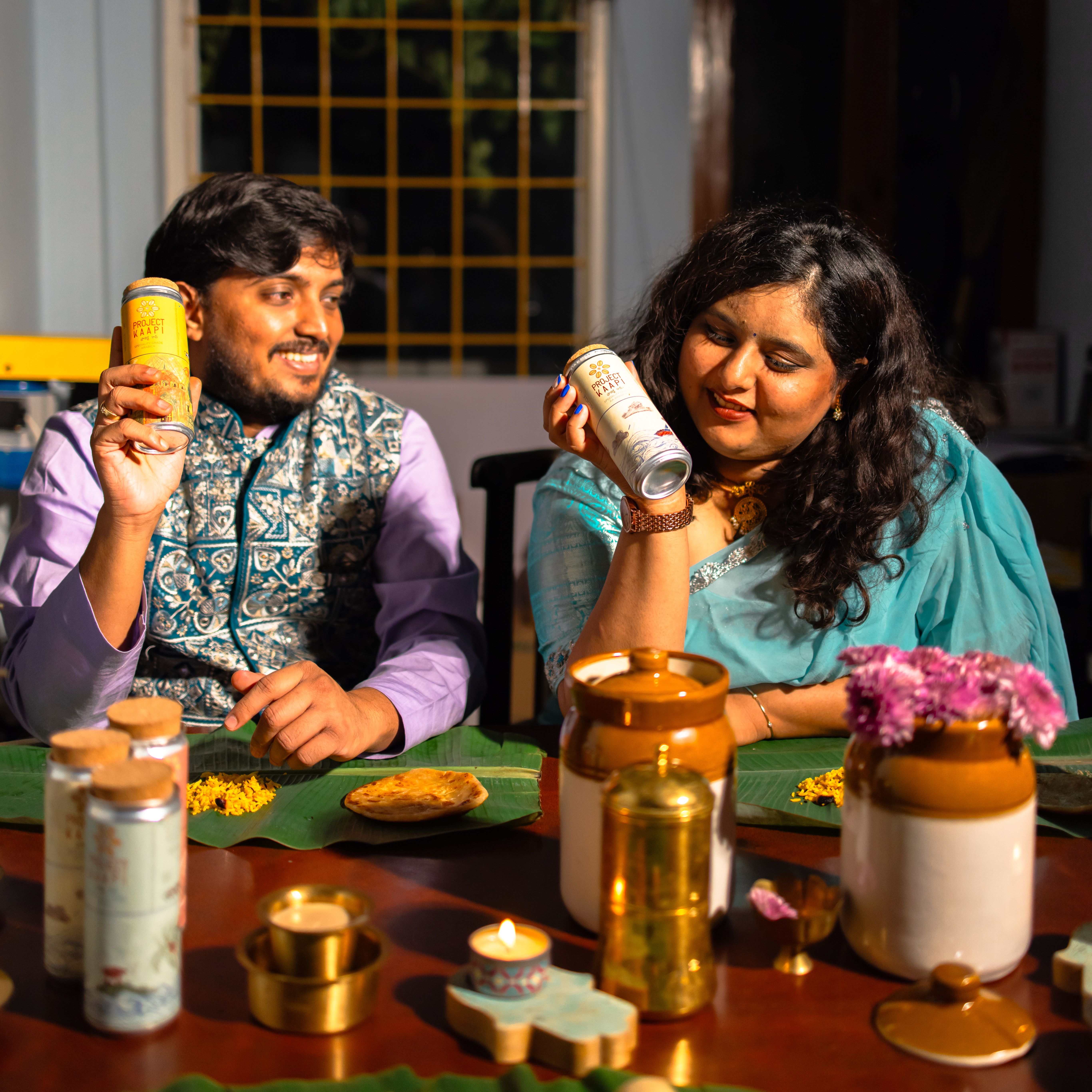 A smiling couple in traditional attire holds up beverage cans at a table set for an Indian meal.