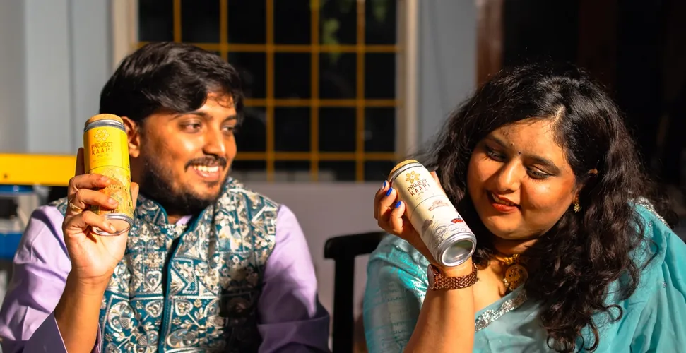 A smiling couple in traditional attire holds up beverage cans at a table set for an Indian meal.