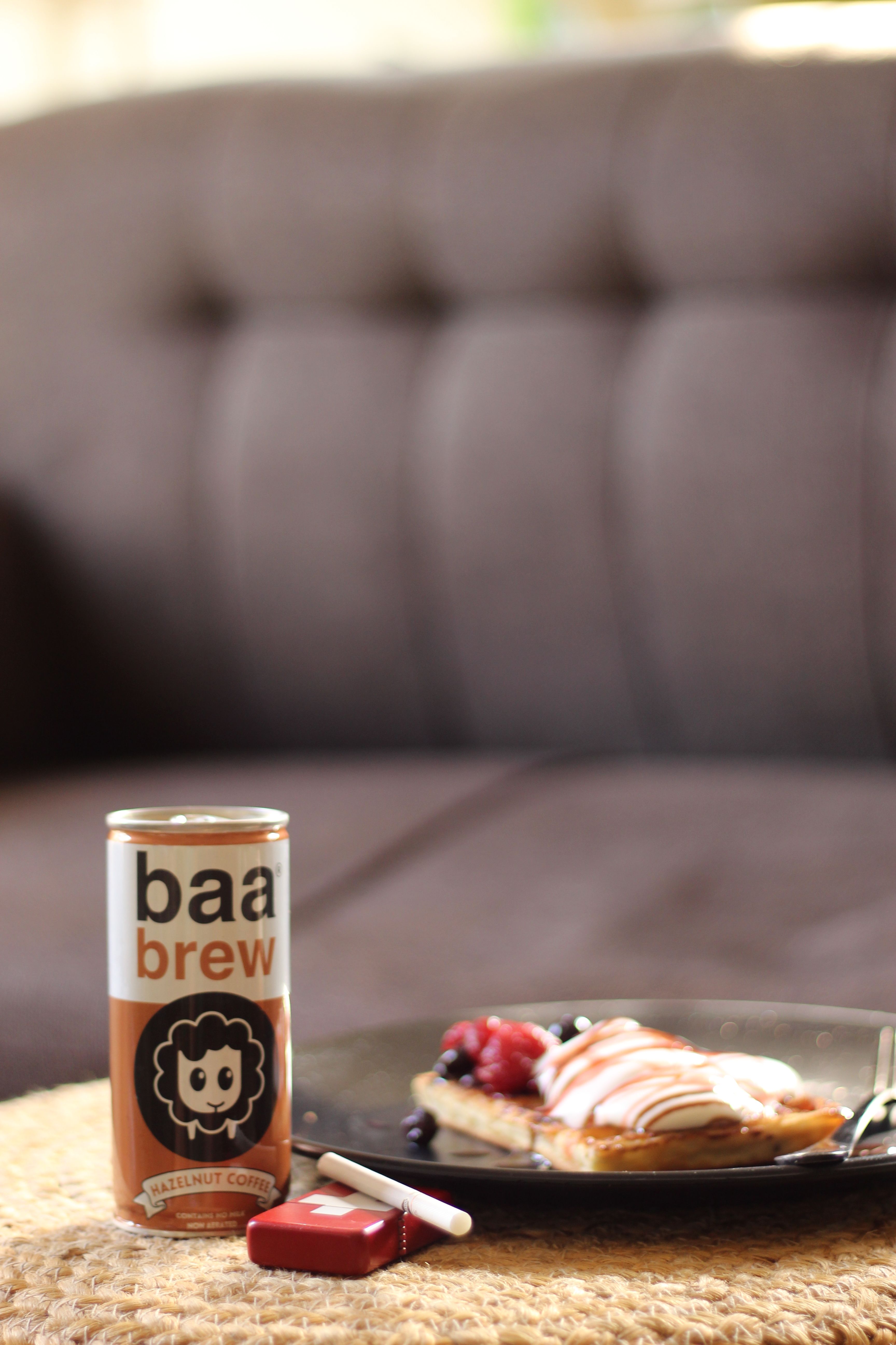 A Baa Brew hazelnut coffee can stands beside a red lighter with a cigarette and a plate of waffles topped with berries and whipped cream, all on a woven mat with a blurred sofa behind.